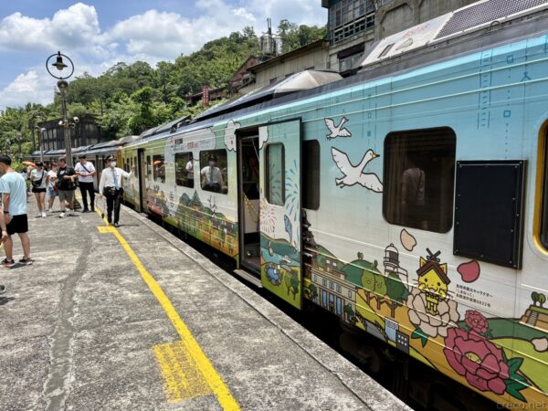 平溪線のホームに停車する観光列車。空色の車体に鳥や町並み、平溪ランタンなどのイラストが描かれた列車が発着し、乗客がホームで待つ。背後は山の緑。