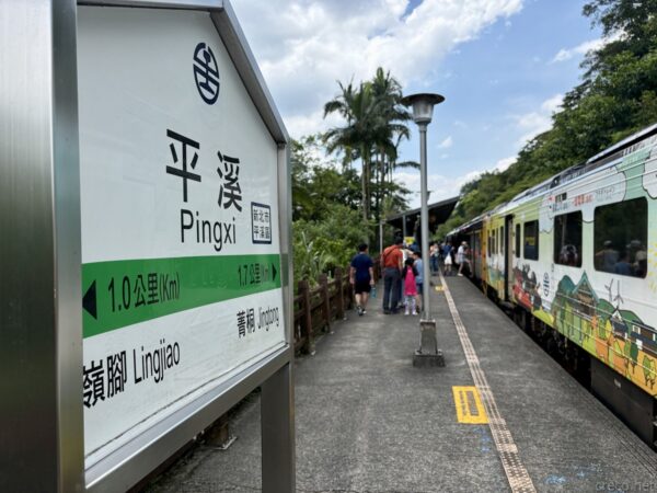 平溪（Pingxi）駅の緑白の駅名標。「菁桐 Jingtong」「嶺腳 Lingjiao」への案内が記され、右に緑ラッピングの列車、椰子の木が立つ。