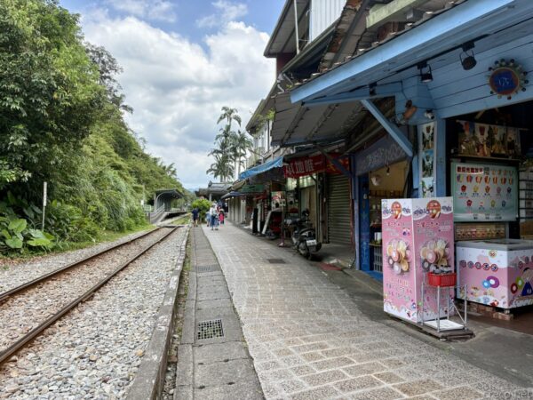 平溪の商店街に沿った線路。片側に軒を連ねる土産物店や飲食店、バイクが停まり、砂利の線路がゆるやかに奥へ延びる。