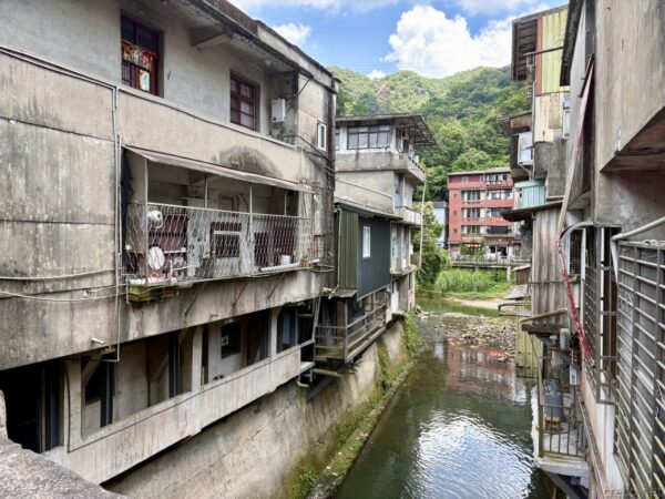 建物に挟まれた小川の眺め。左右に古びた集合住宅がせり出し、ベランダに洗濯物や物が並ぶ。谷底に浅い水が流れ、奥に緑の山。