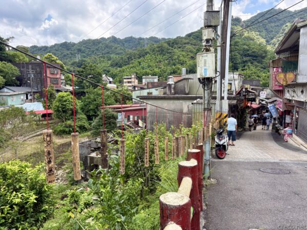 平溪の坂道。赤い天灯を模した飾りが並ぶ柵沿いに家並みが続き、バイクや人影が見える。背後に緑濃い山が迫る山里の路地。