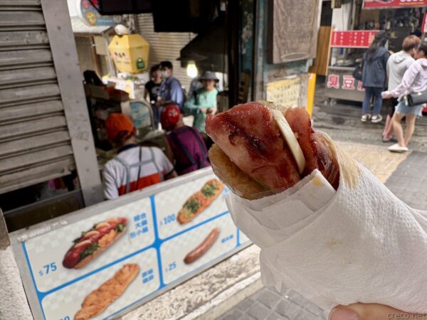 平溪老街の屋台で買った「大腸包小腸」。焼いたもち米の腸詰めに台湾式ソーセージを挟んだB級グルメを紙で包んで手に持つ。背後の屋台には値段付きのメニュー写真が並び、通りには人が行き交う。
