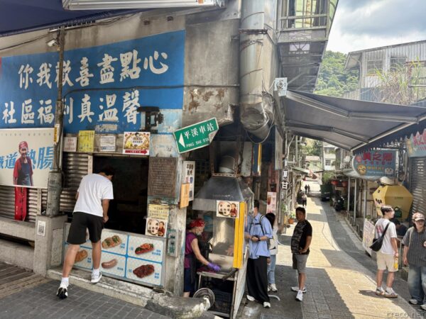 平溪の屋台街の角。青い看板の「鐵道熱腸」ソーセージ店の煙突から煙が上がり、「平溪車站」への緑の案内標識が立つ。客が集まる。