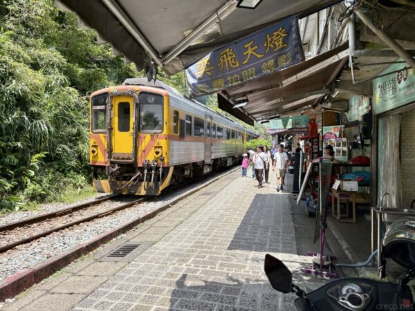 平溪駅の線路脇に停車する橙とグレーの列車。「天燈」の横断幕が下がる屋根の下、線路沿いの土産物店の前を観光客が歩く。