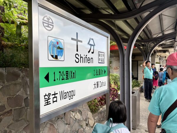 十分（Shifen）駅の緑白の駅名標。天灯を掲げる人物のイラスト付きで、「望古 Wanggu」「大華 Dahua」への案内。水色の服の団体客が写る。