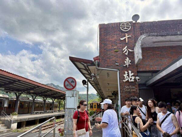 十分車站の赤煉瓦の駅舎正面。「十分車站 Shifen Station」の看板を掲げ、軒下に大勢の観光客が集まる。左にホームと山並みが見える。