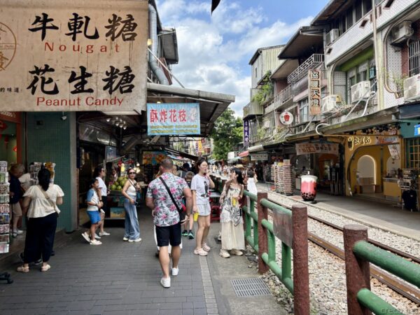 十分老街の賑わい。「牛軋糖 Nougat」「花生糖 Peanut Candy」の看板の下、線路脇の緑の柵の前で観光客が写真を撮っている。