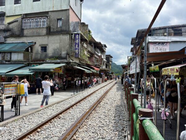 線路がまっすぐ通る十分老街。両側に古い商店が軒を連ね、緑の柵の間を人々が歩く。曇り空の下に山並みが遠く見える。