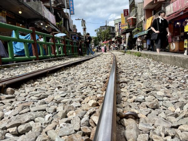 線路に顔を近づけた低い視点の一枚。砂利の間を延びる二本のレールの先に、緑の柵と土産物店、行き交う観光客が写る十分老街。