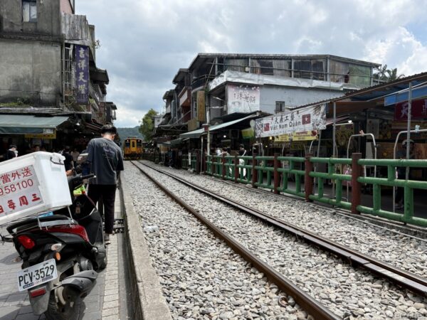 十分老街の線路と商店。奥に橙の列車が停まり、緑の柵沿いに宅配便のバイクや歩く人。「Viet Nam Quan」の看板が右手にある。