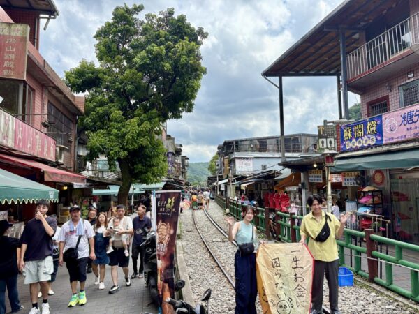 十分老街の中心部。大きな樹木のそばの線路脇で、若い観光客が記念撮影をし、両側に土産物店が軒を連ねる賑やかな通り。