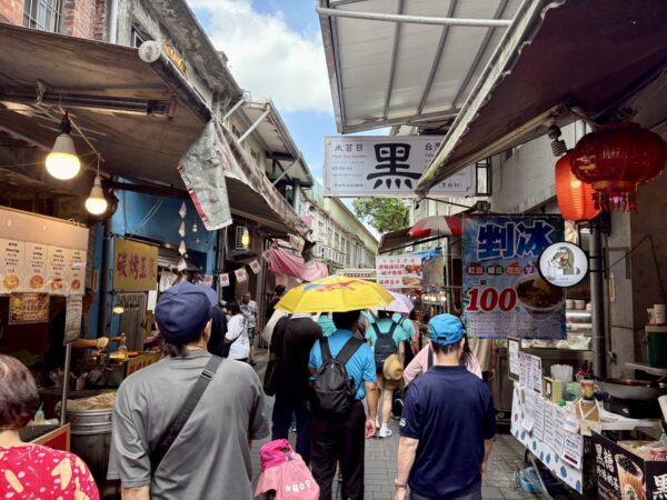 十分の路地の屋台街。「黑」の看板やかき氷「剉冰」の幟が並ぶ細い通りを、黄色い傘を差した団体客が列をなして進む。