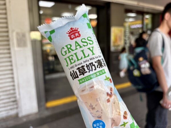 手に持った「仙草奶凍」グラスジェリーミルクのアイスバー。パッケージに商品写真が印刷され、背後にリュックの人と店先がぼやける。