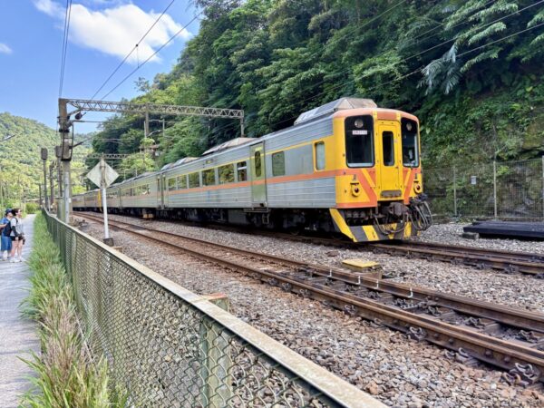 金網フェンス越しに見た走行中の橙とグレーの列車。緑濃い山肌を背に、複線の線路をカーブして通過する電車を捉えた一枚。
