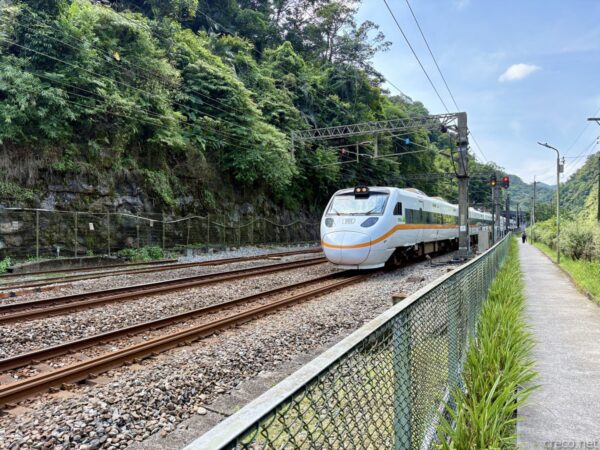 金網越しに見た白い特急型列車。オレンジの帯を巻いた車両が緑の山を背に線路を走り、右奥に信号機と歩道が見える。