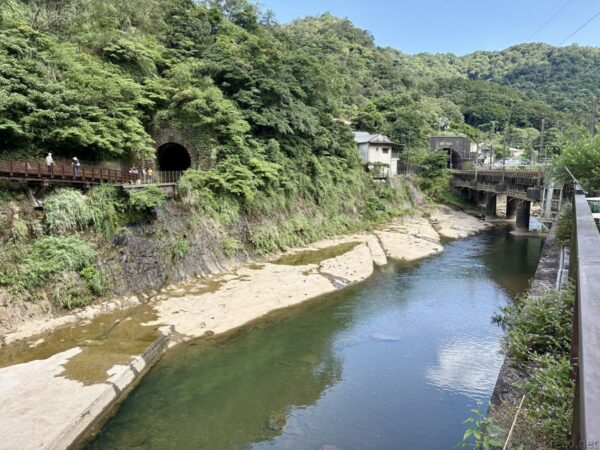 渓谷に架かる橋と山里の家並み。左手に緑の斜面と川、右に鉄橋、正面にトンネルの坑口が見える。深い緑に囲まれた三貂嶺付近の風景。