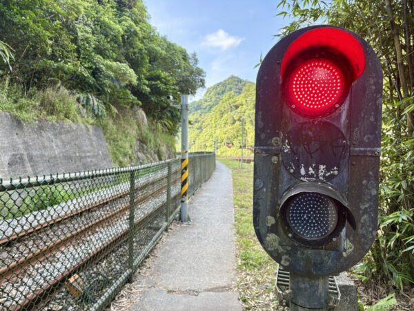 線路脇の歩道に立つ鉄道信号機。赤いランプが点灯し、金網フェンス越しに単線の線路が緑の山へ延びる。青空が広がる。