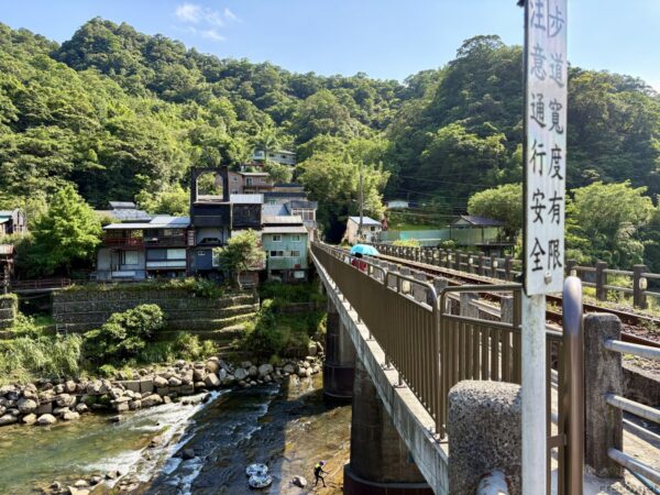 高い位置から見た鉄道橋と山里。橋が谷を渡り、左手に川と古い建物、遠くまで緑の山が続く。手前に注意書きの標柱が立つ。