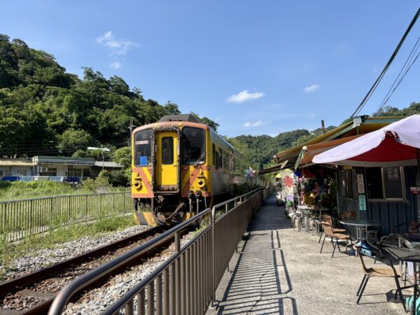 線路脇に停車する黄色い顔の列車。カフェの赤いパラソルとテーブルが右手に並び、緑の山を背にしたのどかな山あいの駅風景。