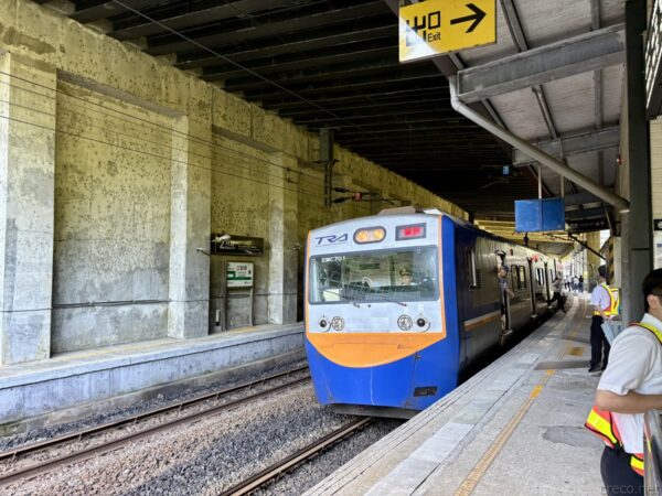 コンクリートの高架下ホームに入線する青と橙の臺鐵列車。「往40 Exit」の黄色い案内が下がり、右に制服の駅員が立つ。