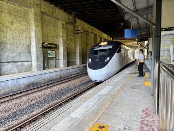 高架下ホームに進入する白い特急型列車。流線形の先頭部が近づき、右に制服の駅員が立つ。コンクリートの薄暗いホーム。
