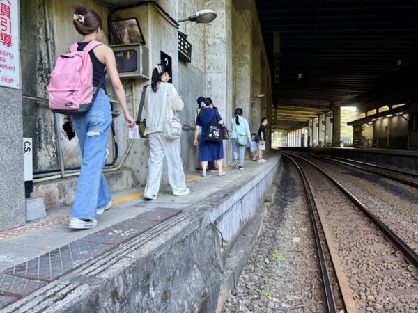低いホームから線路を渡る乗客たち。リュックを背負った人々が古いコンクリートの構造物の脇を歩き、右へ線路が延びる駅構内。