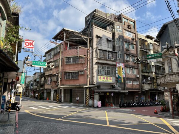 瑞芳老街の街角。赤いタイルや看板の古い低層家屋と数階建てのビルが交差点を囲み、「OK」コンビニの看板やバイク、黄色い区画線の車道が見える。