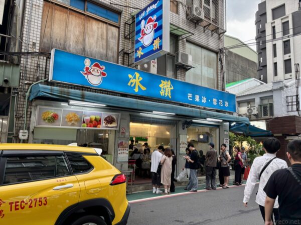 台北の街角のかき氷店「冰讚」。青い看板に「芒果冰・雪花冰」と書かれ、店先に大勢の客が行列する。手前に黄色いタクシーが停まる。