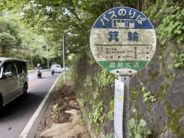 前橋市の赤城山麓、県道沿いに立つ丸い「箕輪」バス停の標識。石積みの擁壁を背にし、道路を白い車とバイク2台が走る。周囲は夏の濃い緑に包まれる。