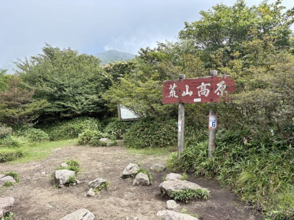 「荒山高原」と記した木製の標識が立つ開けた鞍部。雲に覆われた山肌を背に、地面に丸い石が点在し、6番の標柱も立つ。