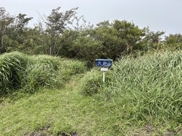 「火起山」の青い山名標が笹原の中に立つ。刈られた草地に踏み跡が続き、低木の茂みと霧に霞む空が広がる稜線上の一点。