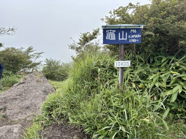 「竈山 Mt KAMADO 1343m」の青い山名標。大きな岩と笹に囲まれ、標柱には標高標も付く。ガスで白く霞む稜線の一角。