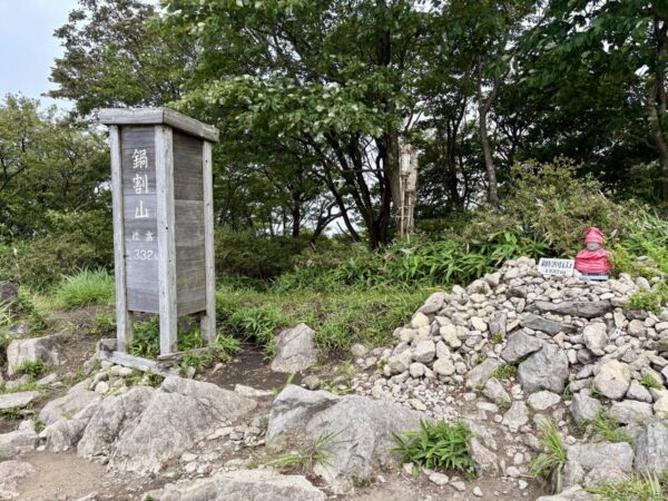 鍋割山の山頂標。「鍋割山 1332」と記した木製の角柱が立ち、傍らに石を積んだケルンと赤い頭巾をかぶった小さな石仏。奥は木立。