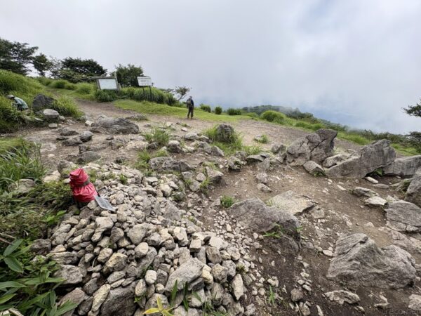鍋割山山頂の岩がちな一角。赤い頭巾の石仏をのせたケルンと登山者、案内板が立ち、その先はガスに沈む谷へと落ち込む。