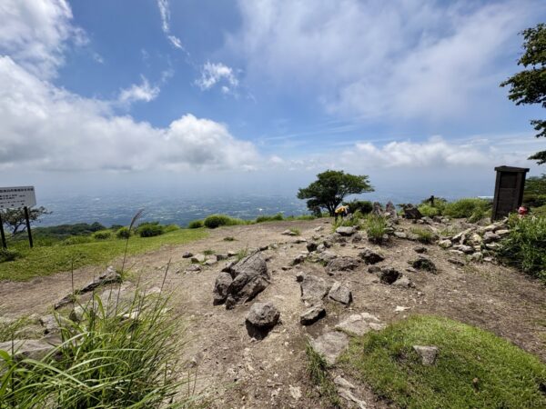 鍋割山山頂の広場。岩混じりの裸地に案内板や木の標識、花を供えたケルンが並び、青空の下に関東平野が霞んで見える。