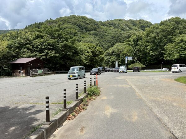 姫百合駐車場に下山。木造の建物が立つ舗装された駐車場に数台の車が停まり、背後は緑の茂る山、車止めのポールが並ぶ。